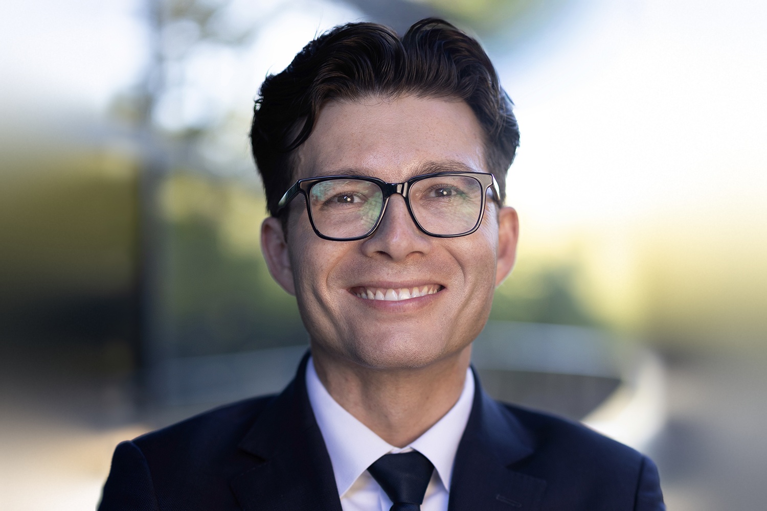 Headshot of Sergiu Pasca smiling, wearing dark-framed glasses, a navy suit, white shirt and dark tie, in front of a softly blurred outdoor background.