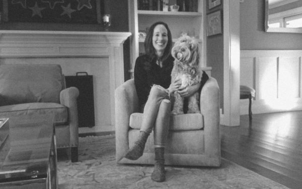 Amy Zegart smiling, sitting in a chair with her dog, whose head is cocked to the side