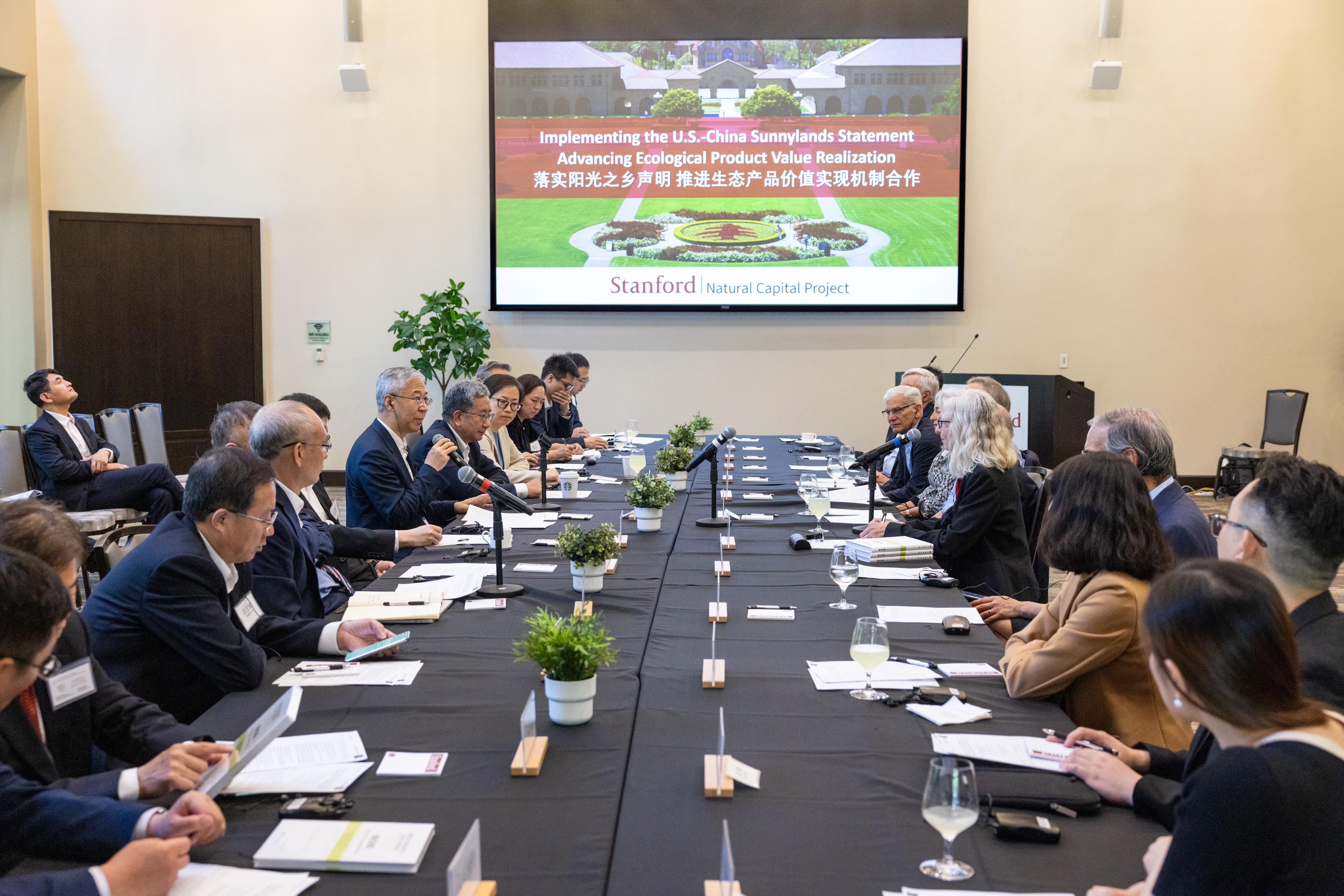 People sitting at a long conference table with microphones. In front of them a screen shows a presentation titled Implementing the US-China Sunnylands Statement, Advancing Ecological Product Value Realization 
