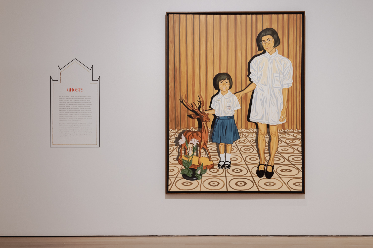 A painting of an adult woman and a child standing together hangs on the gallery wall beside a plaque. The text is too small to read, but the title reads, "GHOSTS".