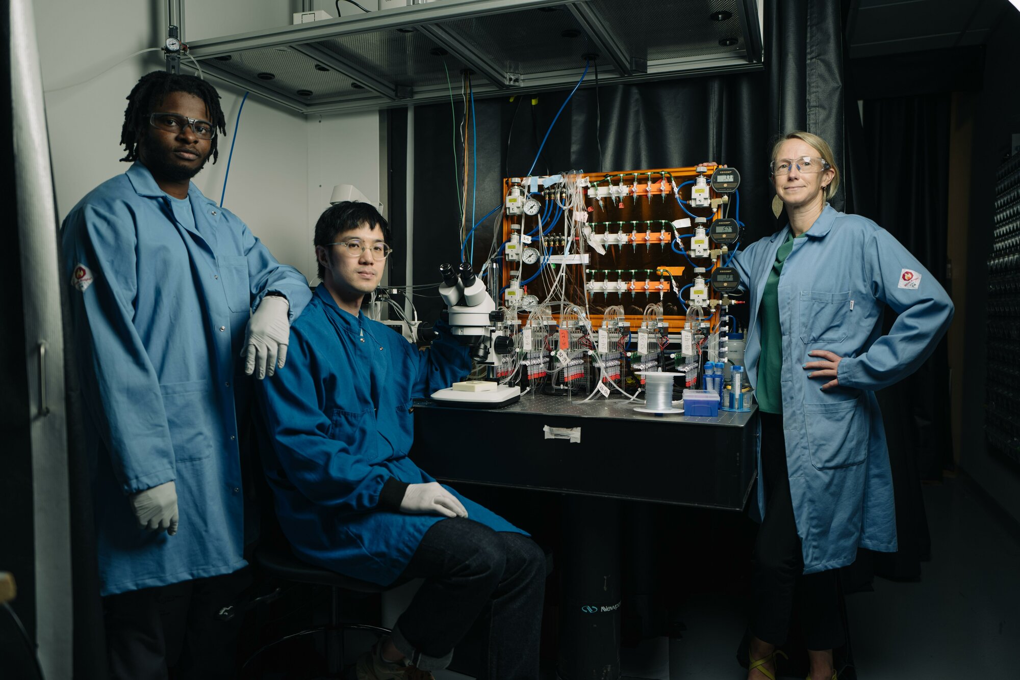 Three researchers in lab coats standing by equipment.