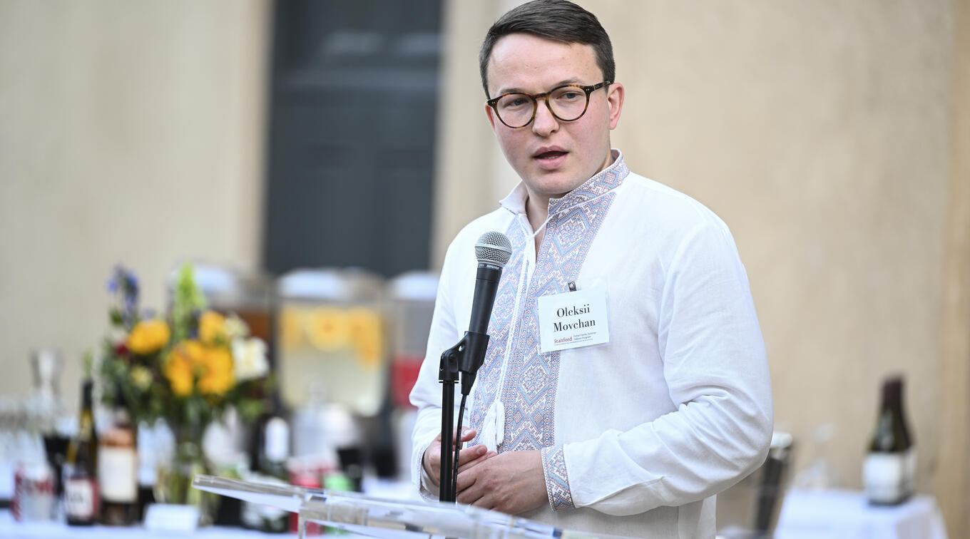 Oleksii Movchan in traditional attire stands at a podium with a microphone, addressing an audience at an event.