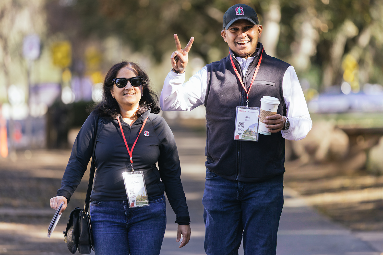 Two attendees walk along a path, one making a peace sign while holding a coffee cup, each wearing a name badge.