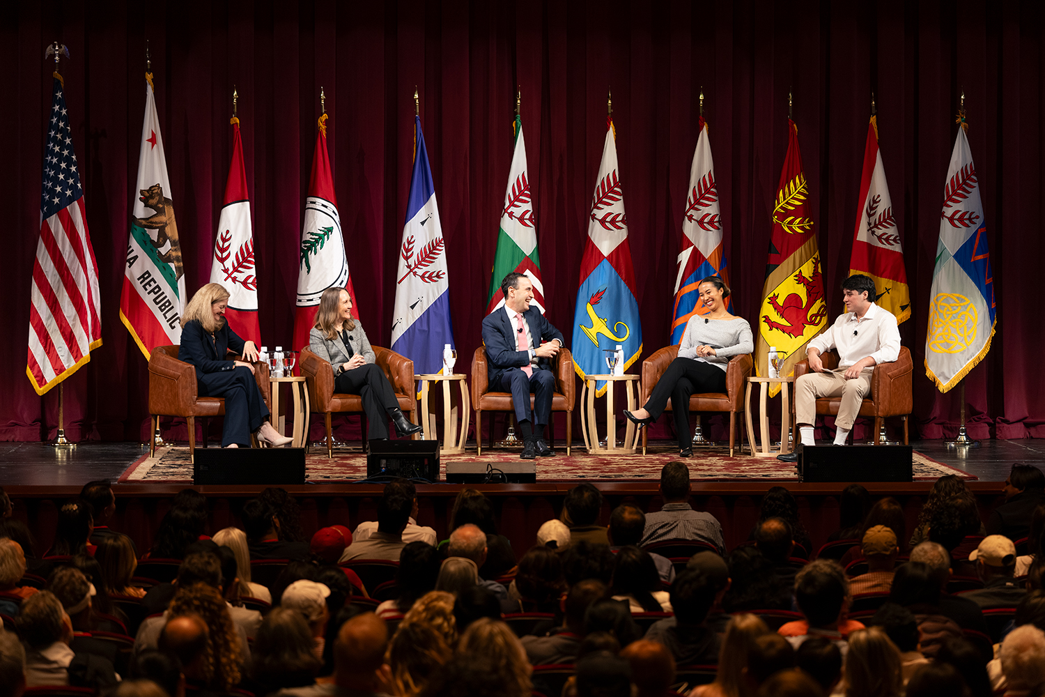 A panel discussion features five speakers seated on stage, with various flags displayed behind them and an audience visible in the foreground.