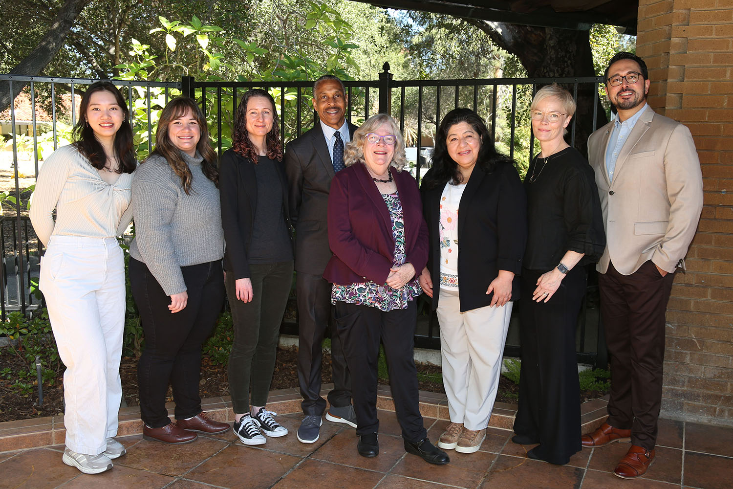 Group of eight people posing outdoors by a wrought-iron fence and brick wall.