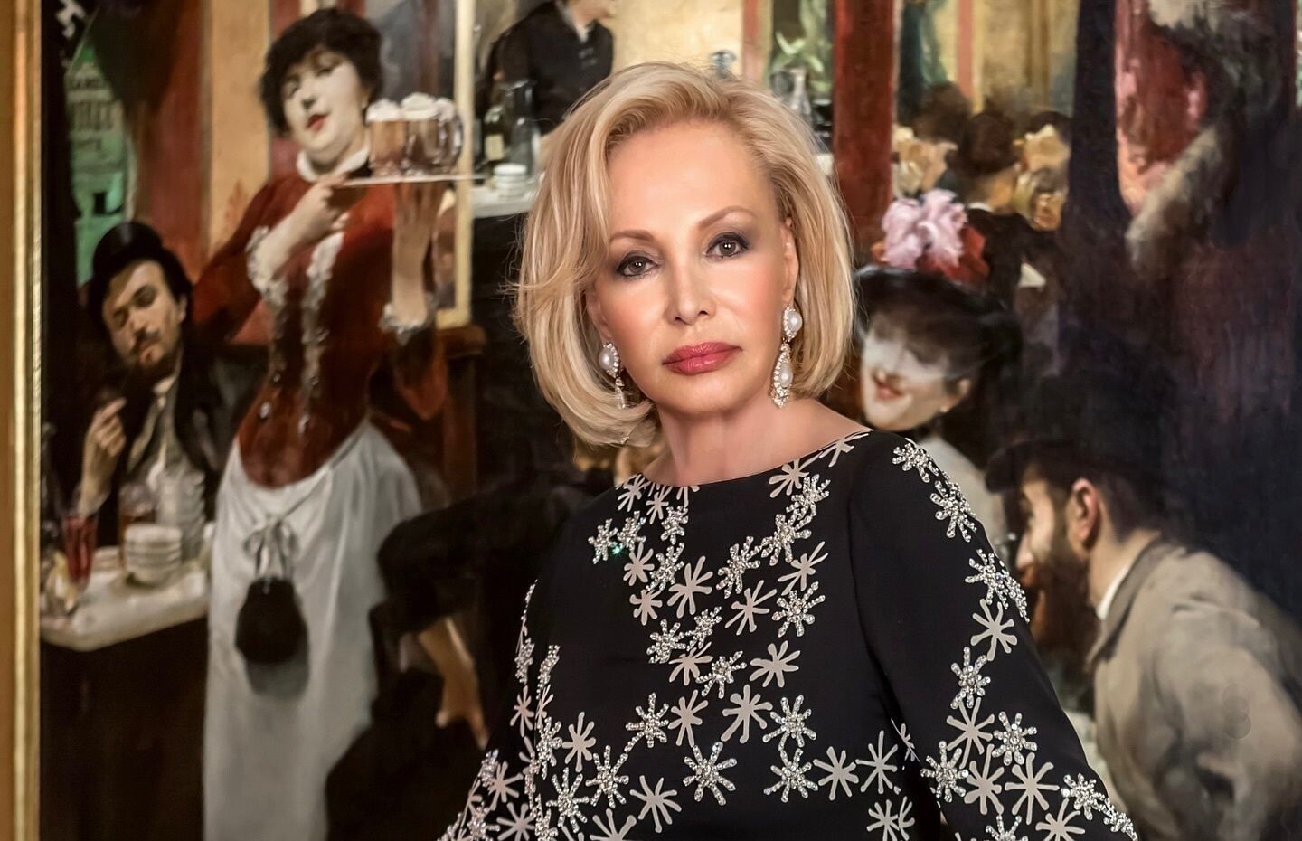 Iris Cantor in a sparkly black dress stands in front of a vibrant painting depicting a bar scene.