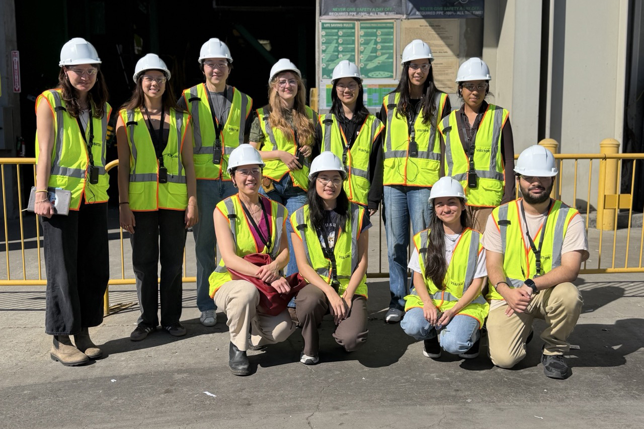 Group of eleven people wearing white hard hats and bright yellow safety vests pose together at a construction site.