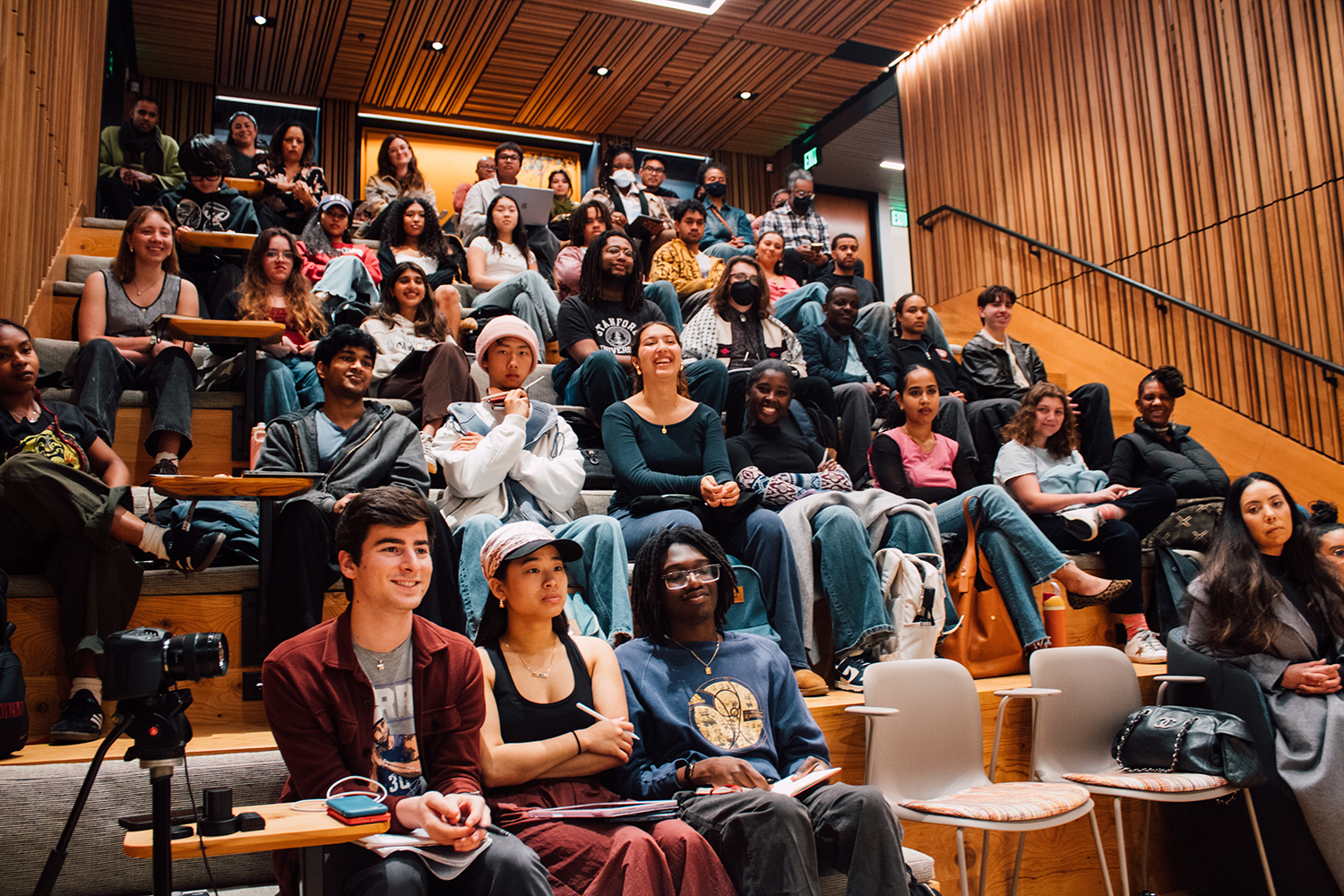 An audience fills a tiered seating area, attentively watching a presentation while some take notes.