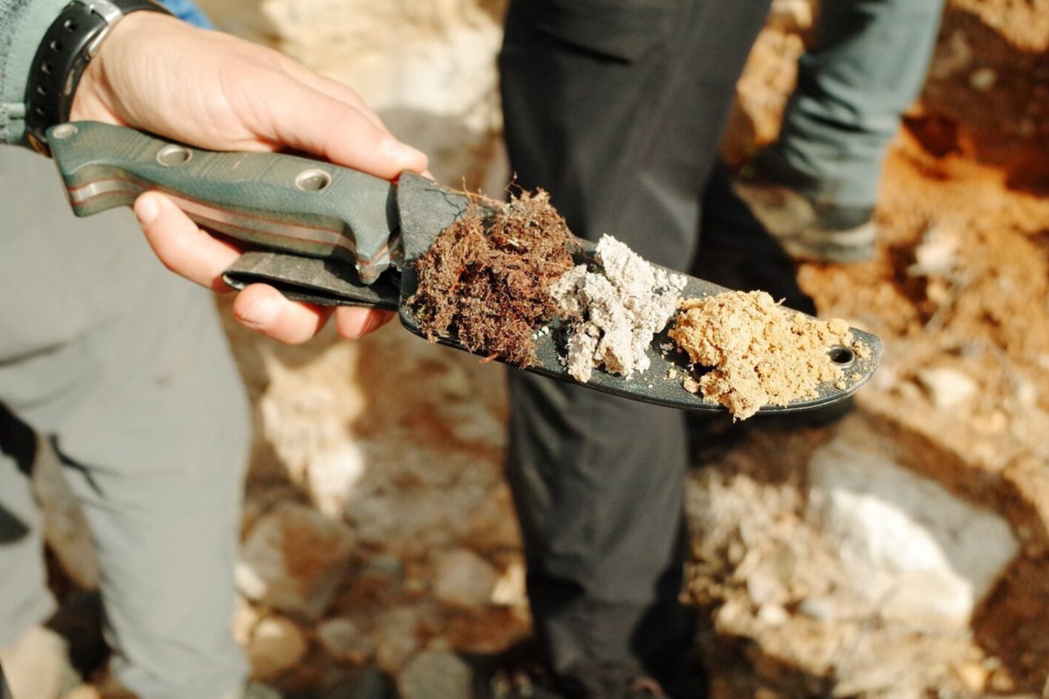A person holds a knife displaying different soil samples.