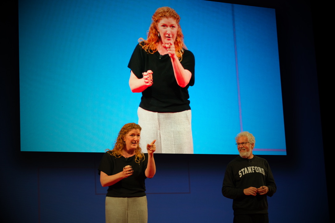 Lisa Rowland in black blouse gestures while presenting on stage alongside Dan Klein in Stanford sweatshirt with large screen behind them.
