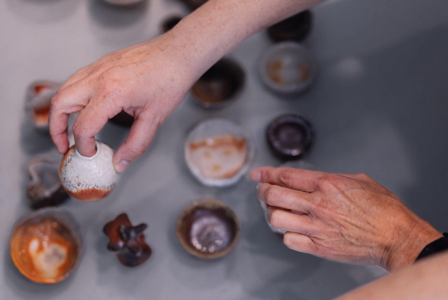Hands select handmade ceramic bowls with various glazes displayed on a table.