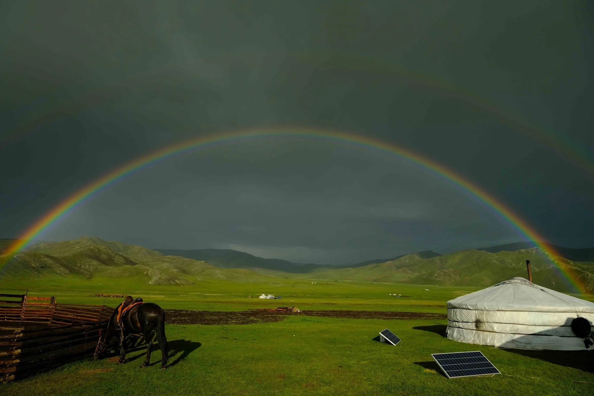 A rainbow arches over a green landscape with a black horse near a yurt and solar panels, under a dark, cloudy sky.