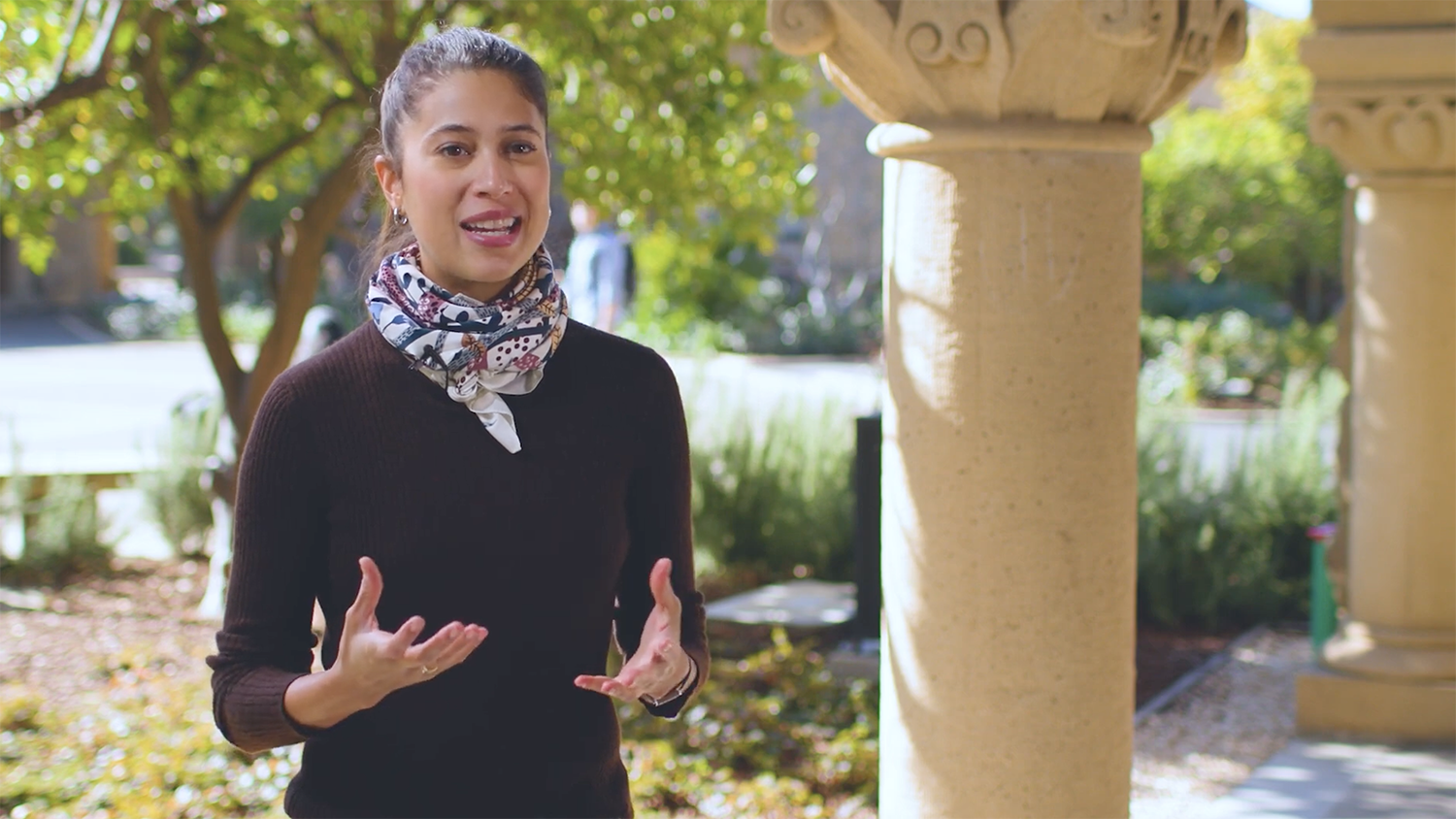 Beatriz Stix-Brunell speaks outdoors near a stone column, gesturing as they explain something in a natural setting.