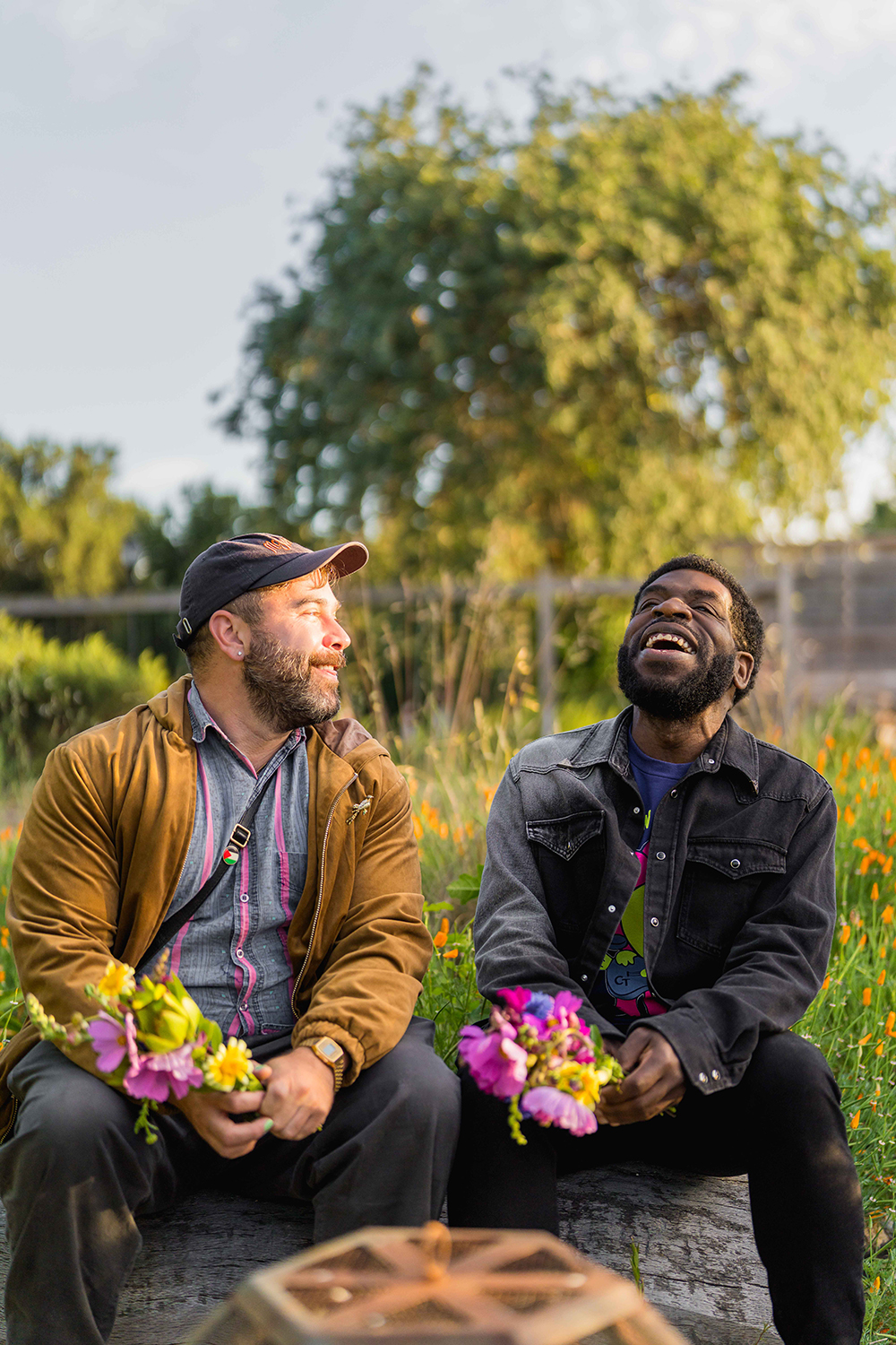 Hanif Abdurraqib and Sam Sax sit outside in a flower-filled garden, each holding colorful bouquets after a floral arrangement workshop.