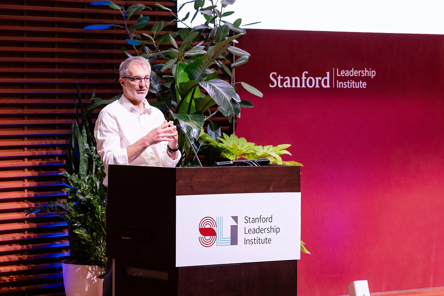 Ken Shotts gestures while presenting at the Stanford Leadership Institute, surrounded by plants and a branded backdrop.