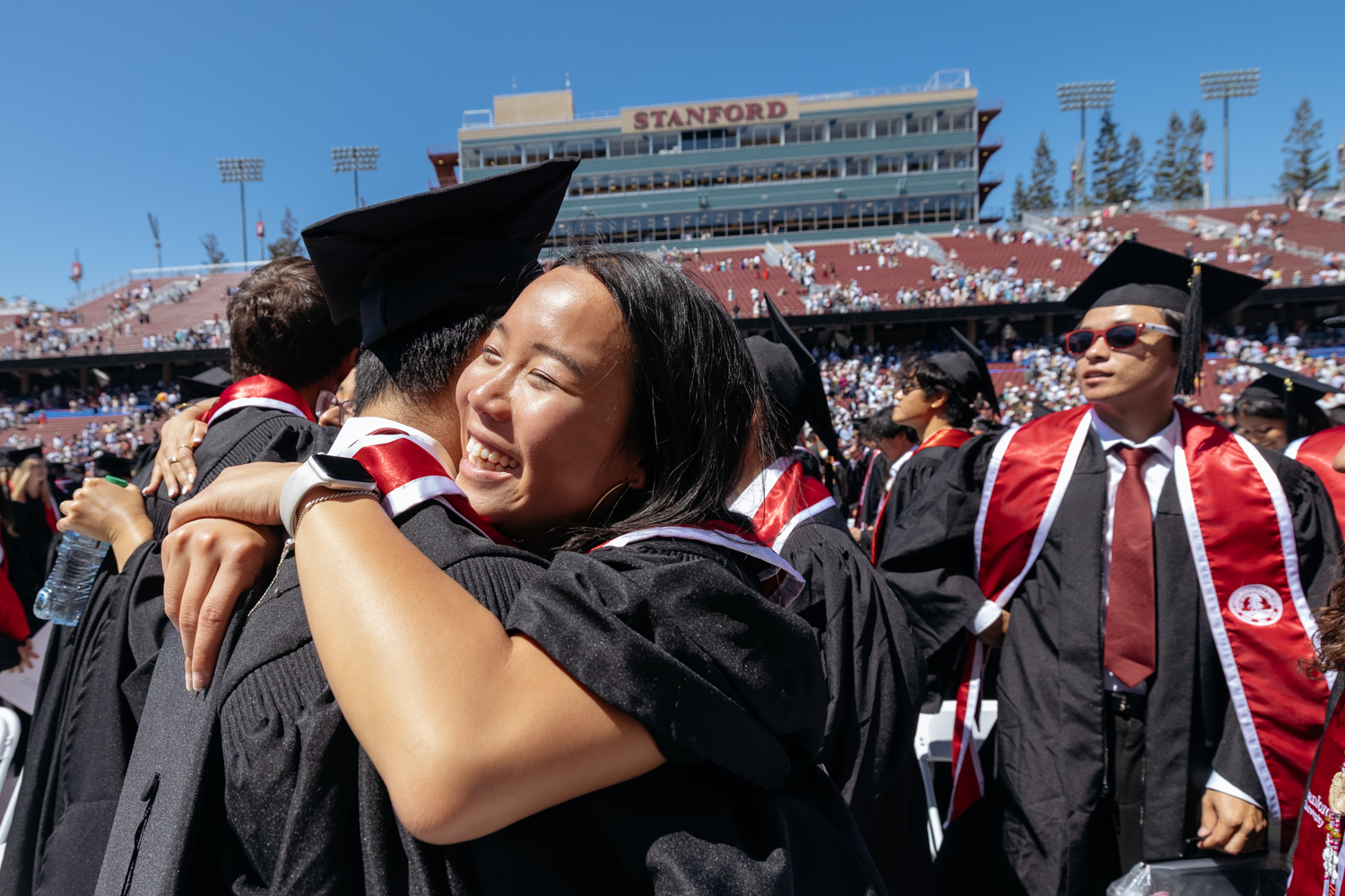 Two graduates in caps and gowns embrace during commencement ceremony at stadium.