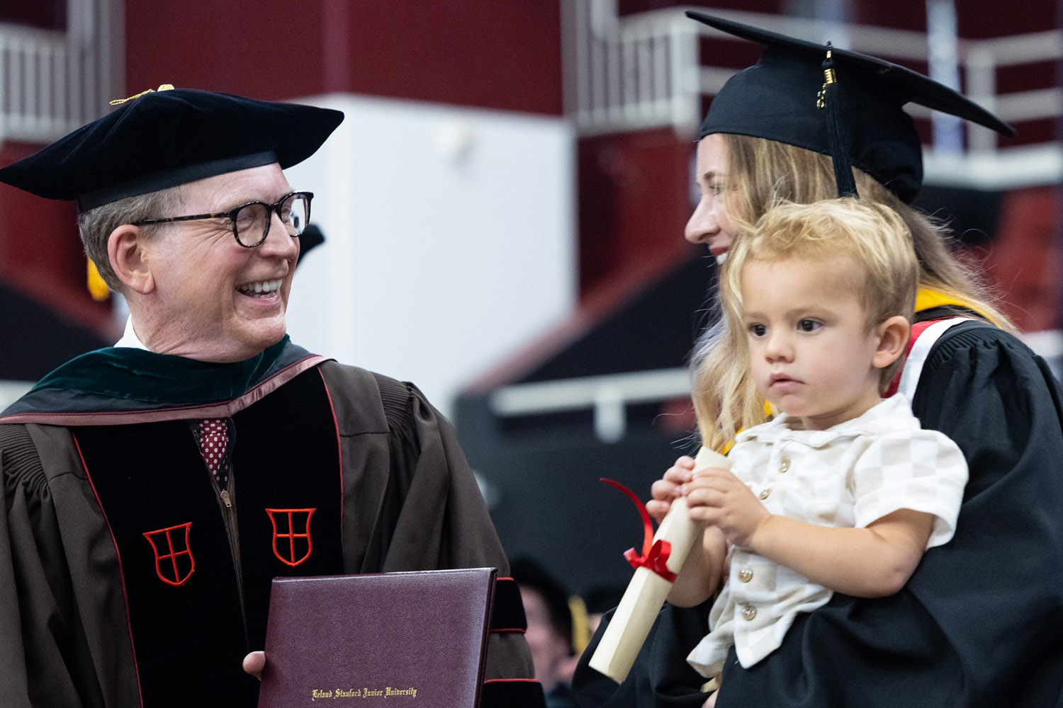 Dean presents diploma to graduate in cap and gown holding young child.