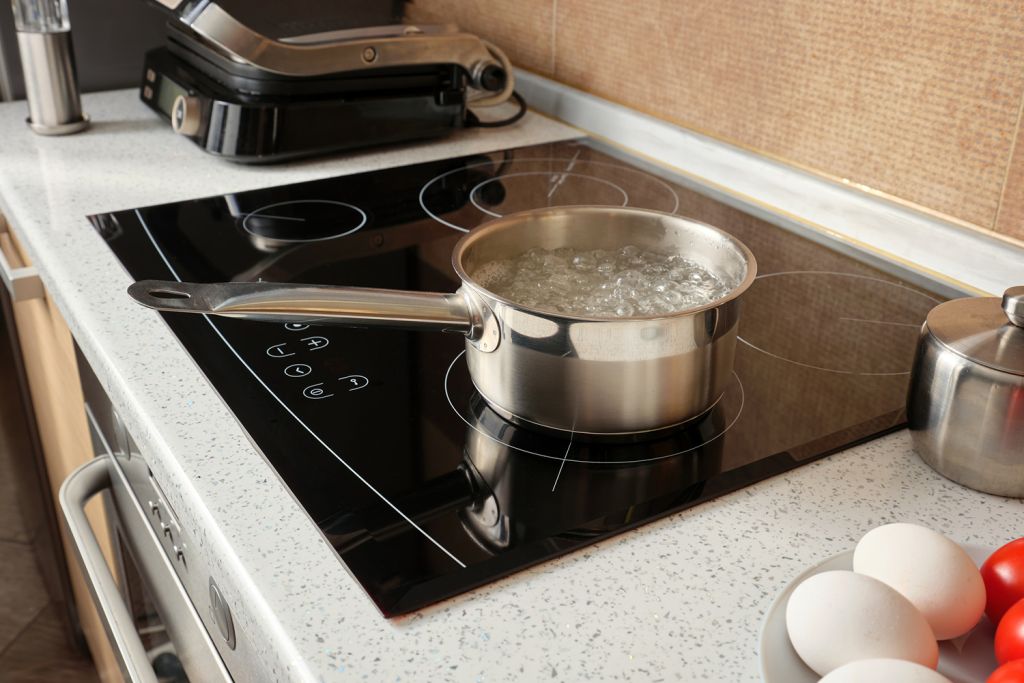 A metal pot filled with boiling water sits on a modern stovetop near eggs and tomatoes on a plate.
