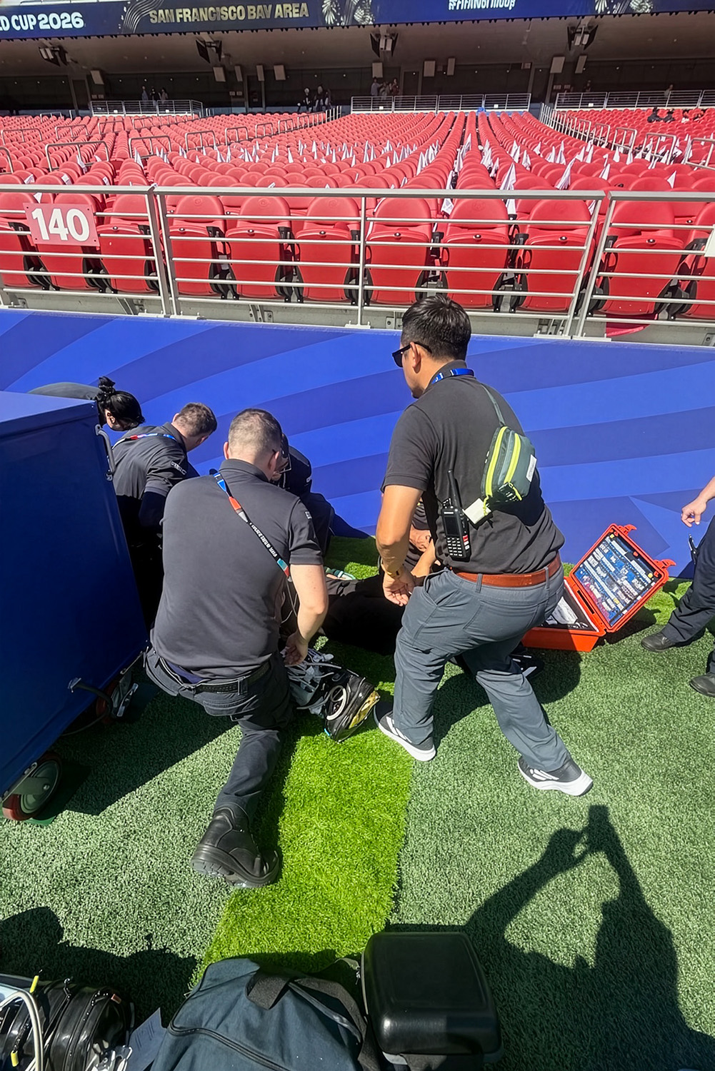 Medical staff kneel to treat someone during a medical response simulation on a stadium field while a colleague stands nearby with a radio and open emergency kit.