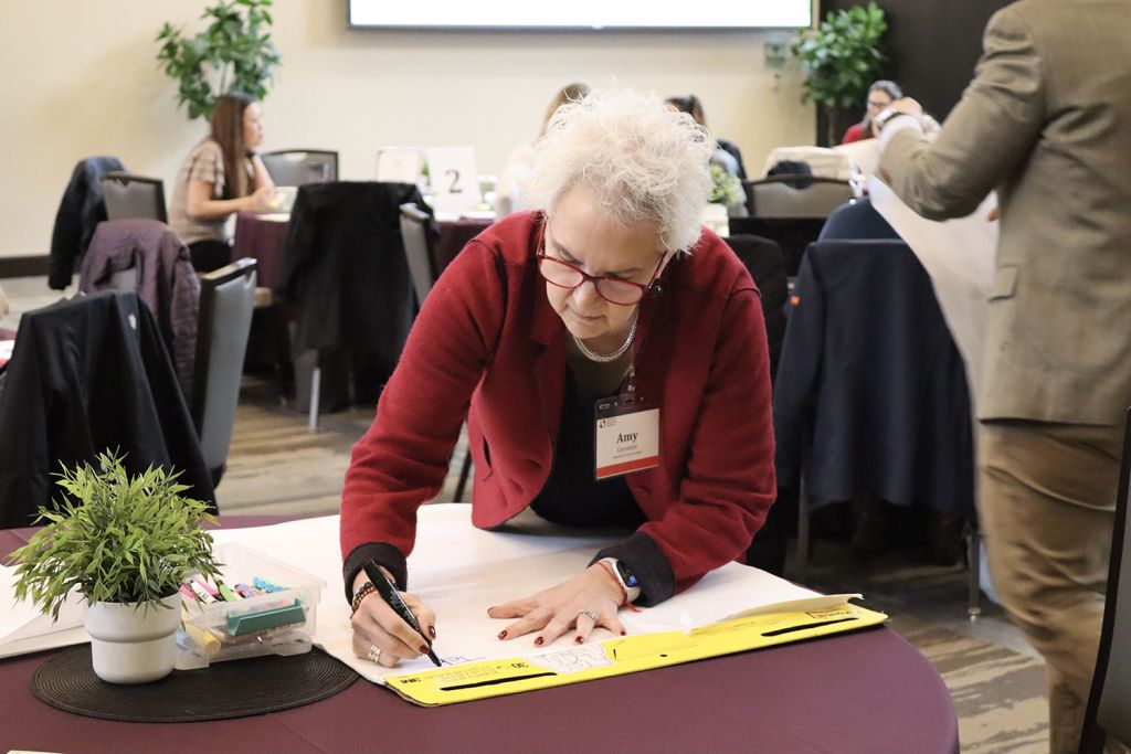 A woman with curly gray hair writes on a large sheet of paper at a round table during a meeting, surrounded by other participants.