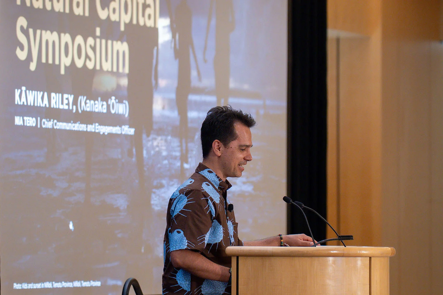 A man in a blue patterned shirt speaks at a podium during the Natural Capital Symposium, with a presentation screen behind him.