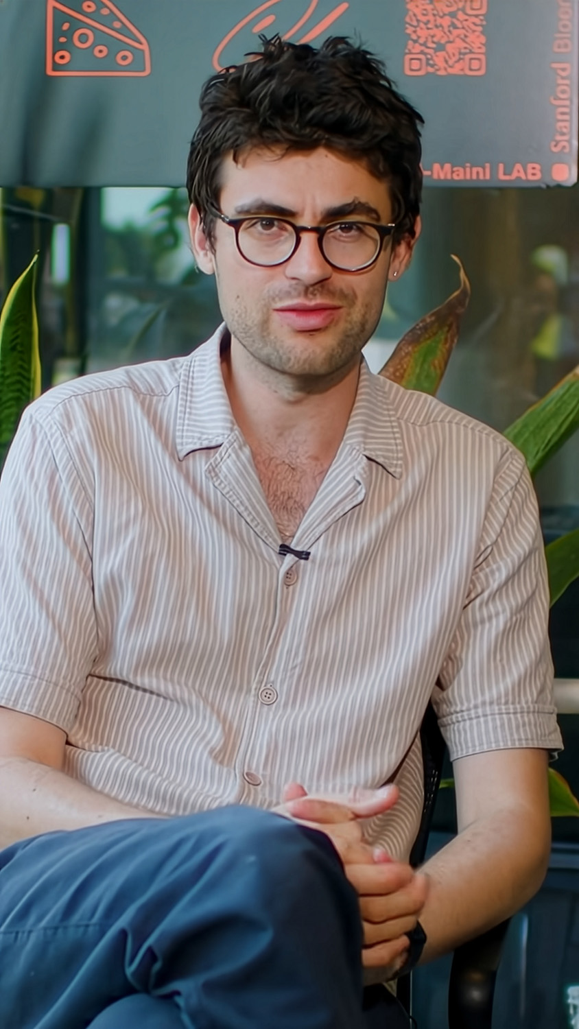 Vayu Hill-Maini in glasses and a striped shirt sits and speaks to the camera during an interview, with lab signage and plants behind him.