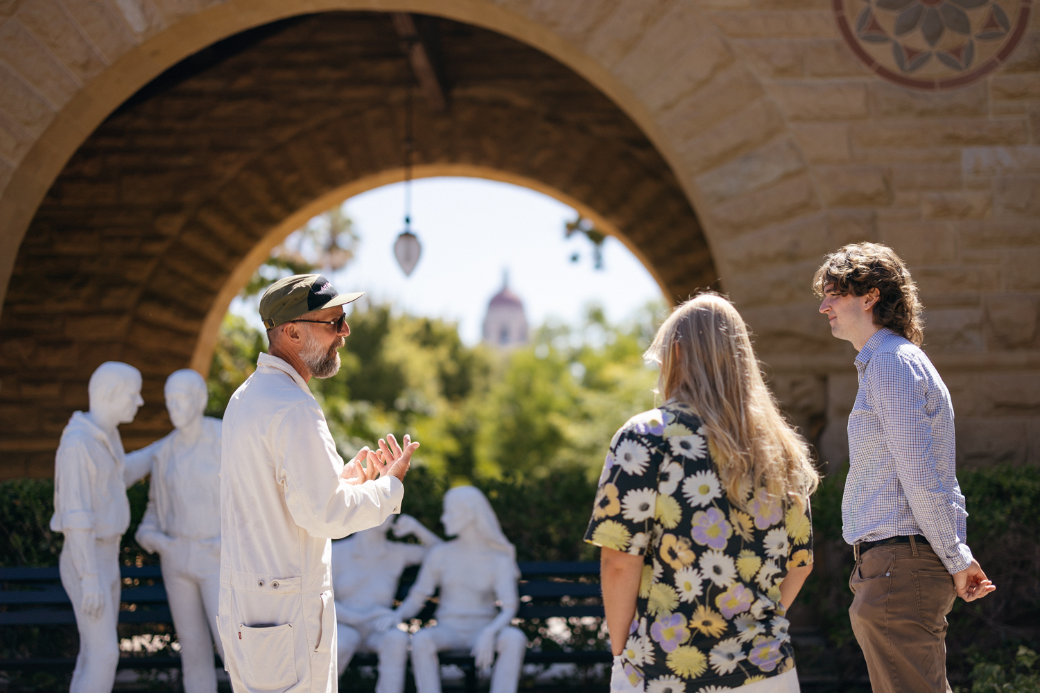 Mark Shunney gestures while speaking to two visitors beside sculpture of white figures under a stone arch.