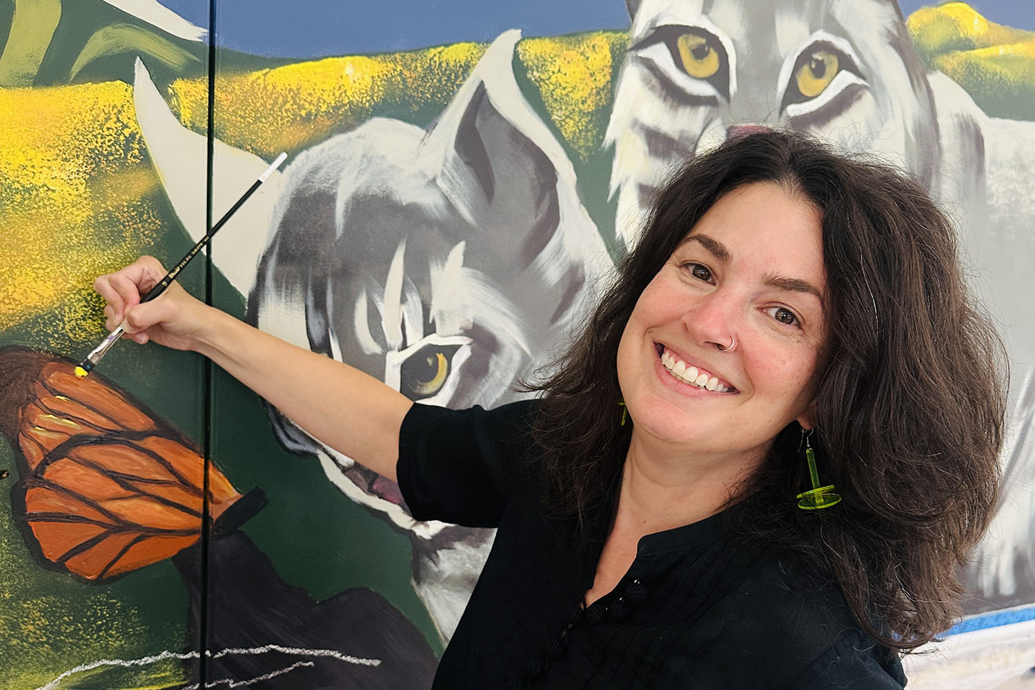 A smiling woman holds a paintbrush near a monarch butterfly she painted on a mural featuring bobcats.