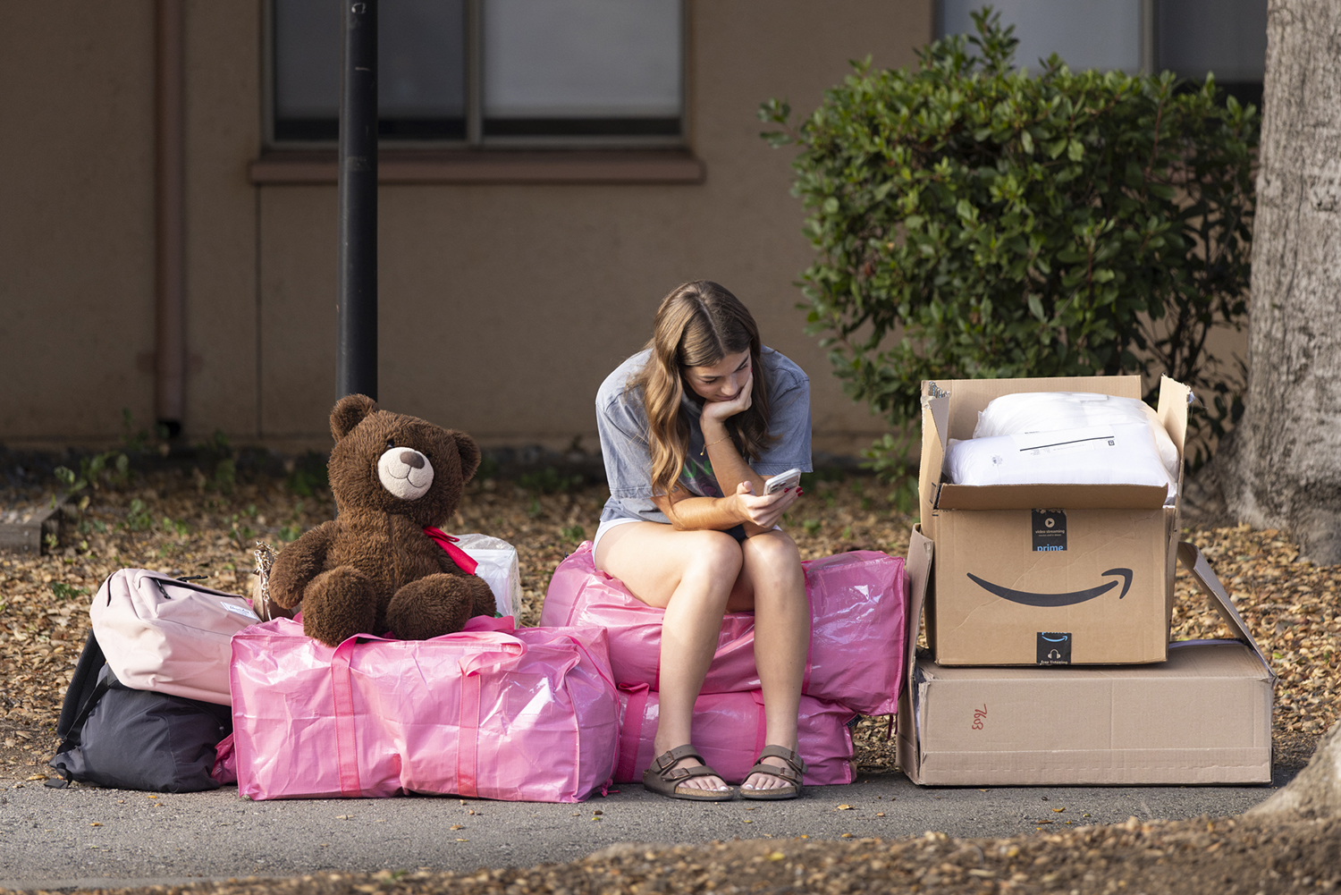 student sits on a pile of moving bags and boxes outside her dorm while checking her phone.