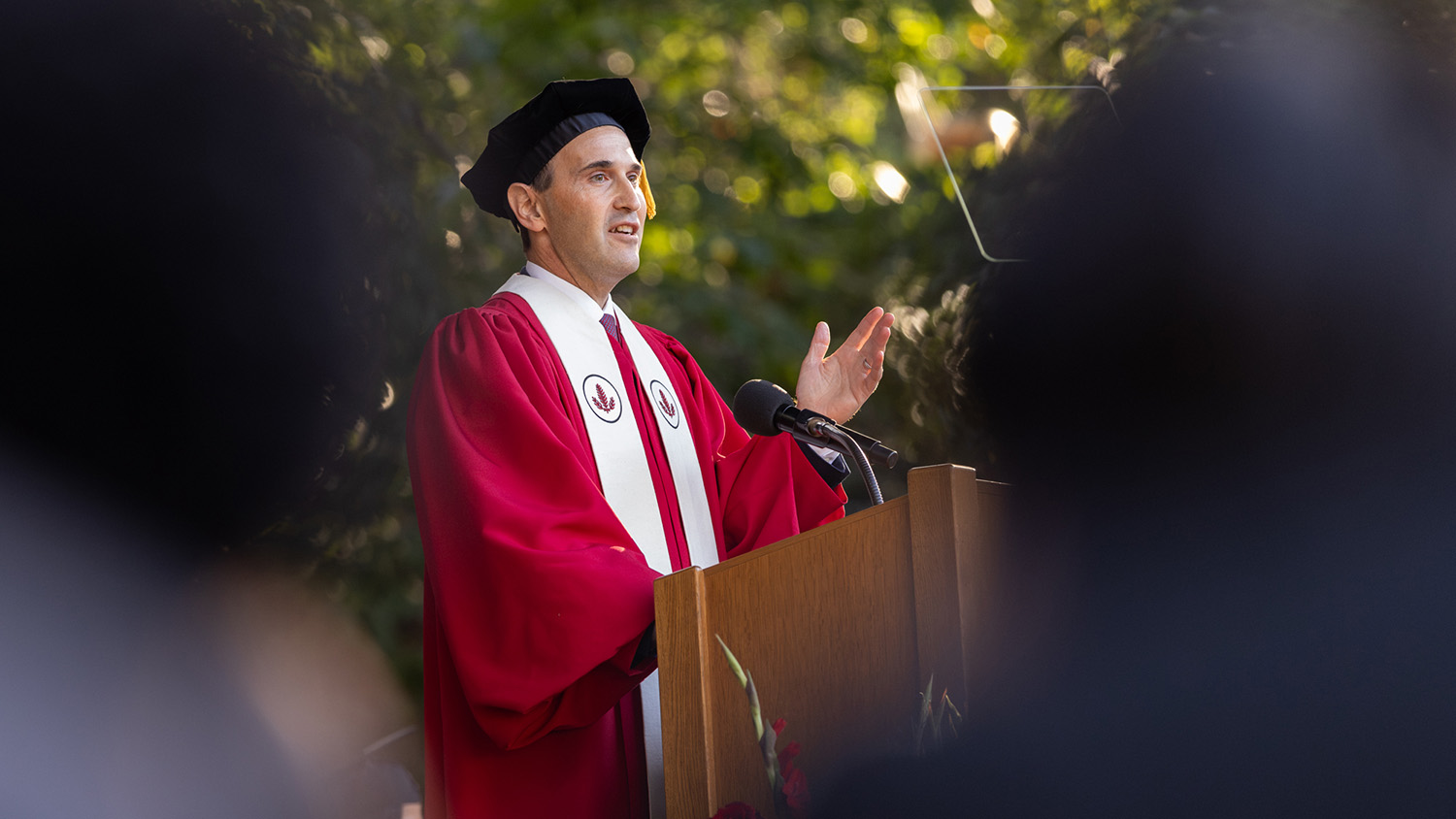 President Levin in red academic regalia and speaking at a podium outdoors.