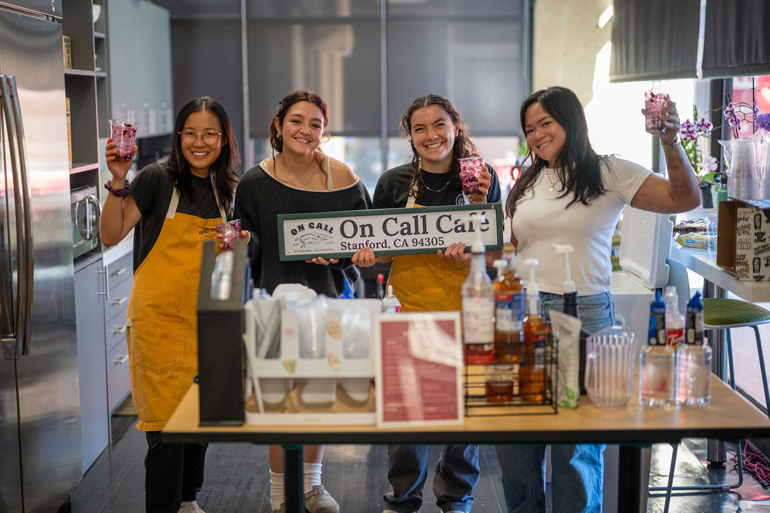 Four baristas hold up drinks while proudly displaying a sign for their student-run café.