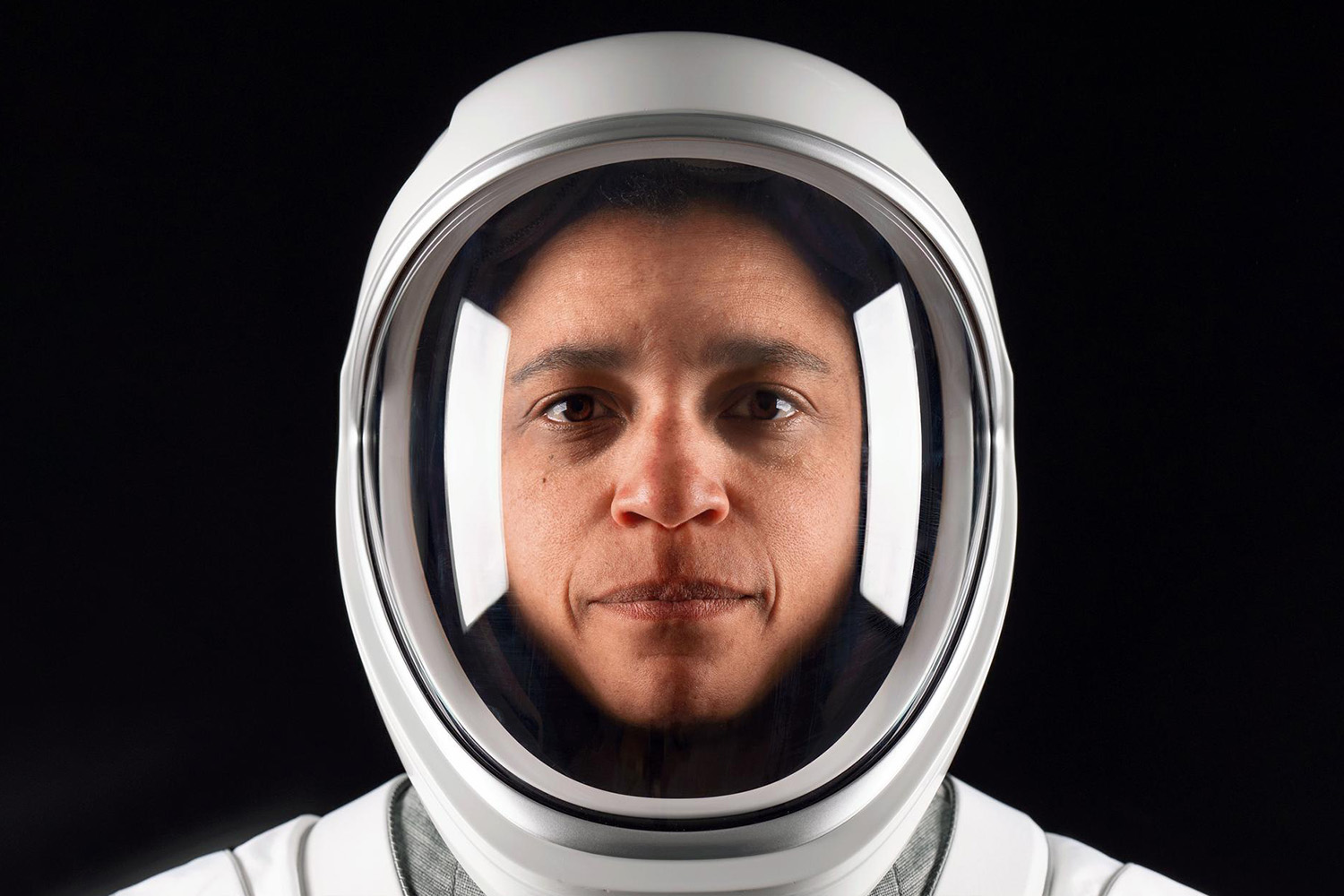 Close-up portrait of Jessica Watkins in a white spacesuit and helmet with the visor down, looking at the camera against a black background.