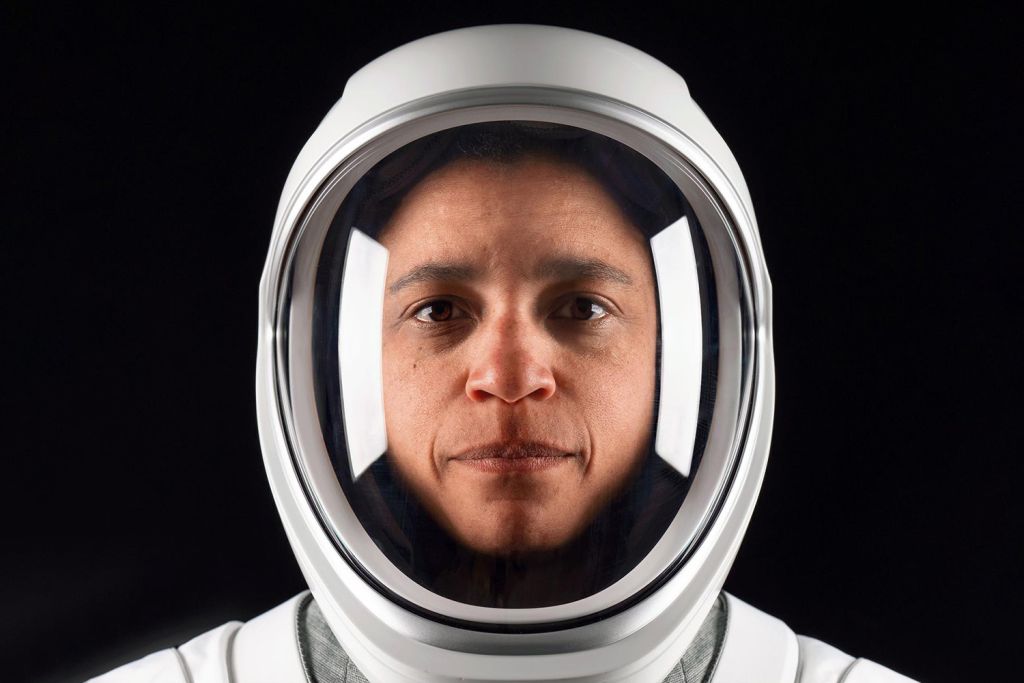 Close-up portrait of Jessica Watkins in a white spacesuit and helmet with the visor down, looking at the camera against a black background.