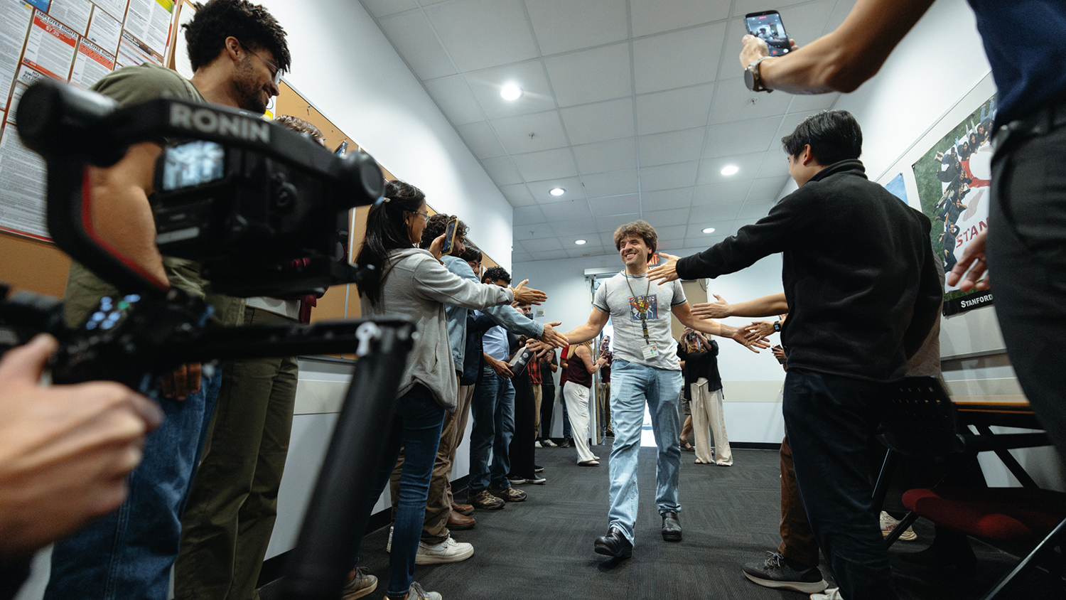 Karl Deisseroth walks down a hallway lined with people who reach out to shake his hand, while others film on phones and a camera.