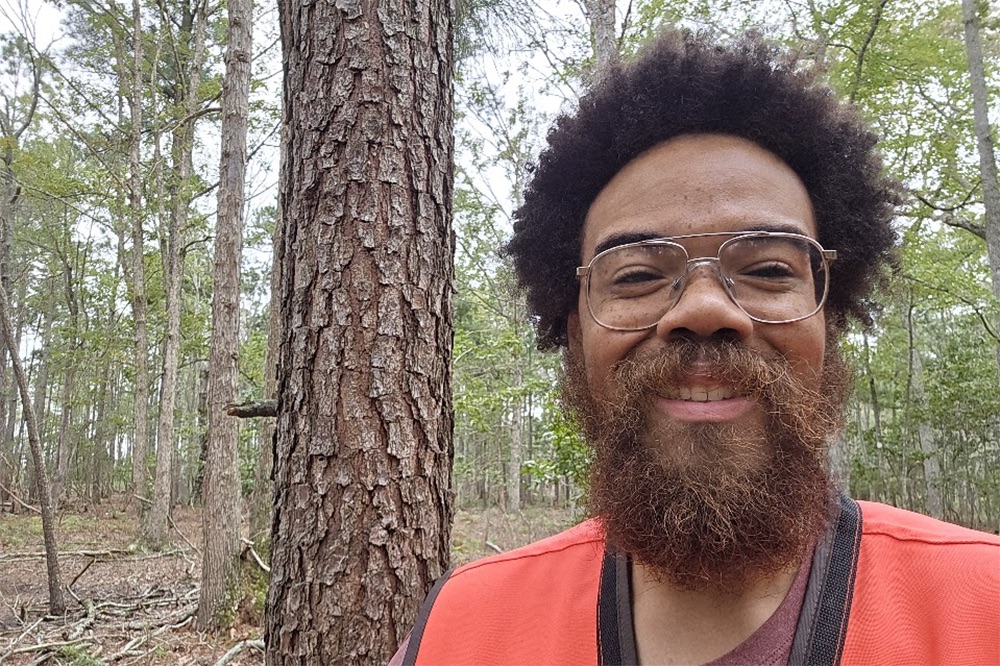 A smiling man in glasses and an orange vest stands beside a pine trunk in a forest.