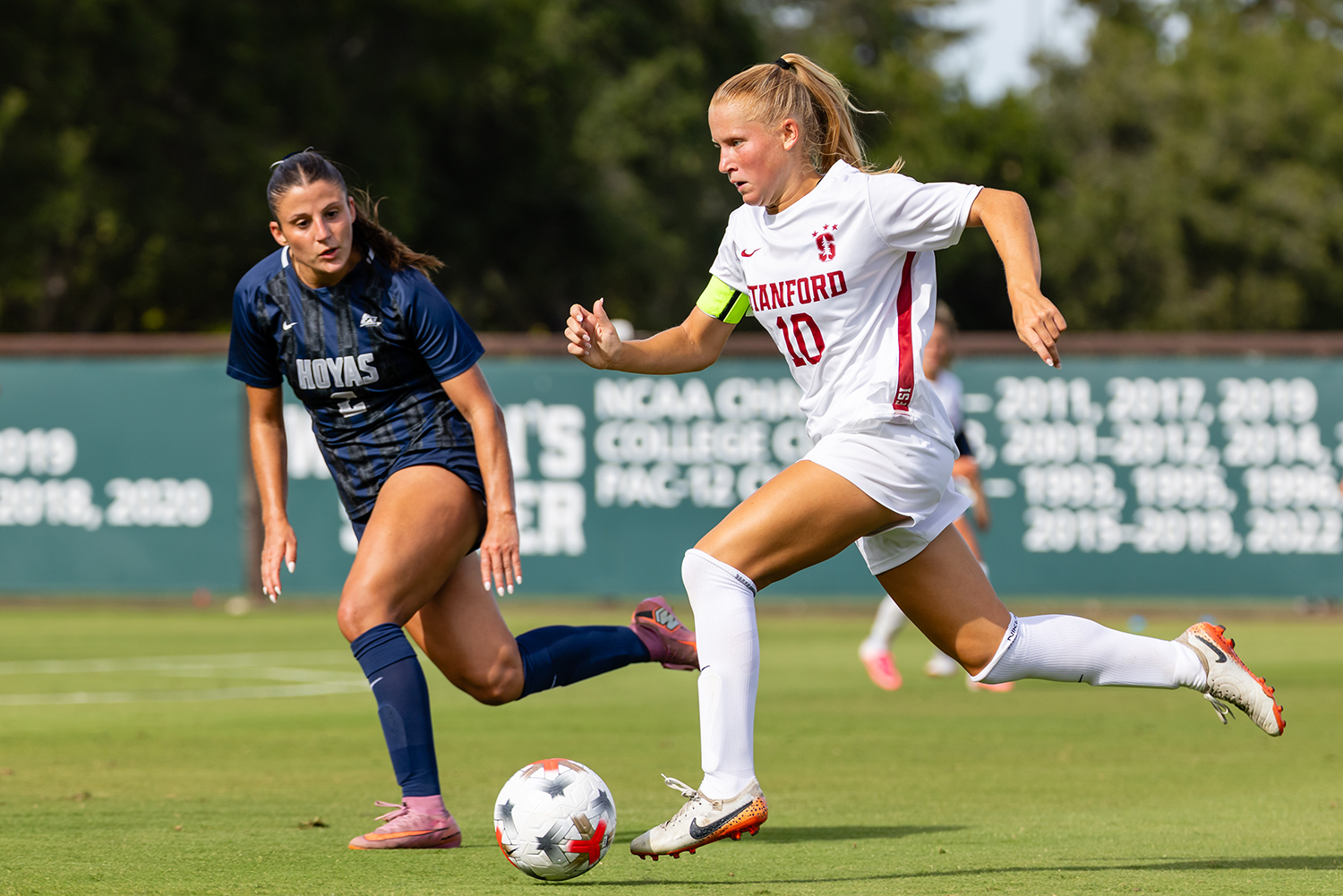 A Cardinal soccer player in a white jersey with a captain’s armband dribbles the ball past a Georgetown defender in navy.