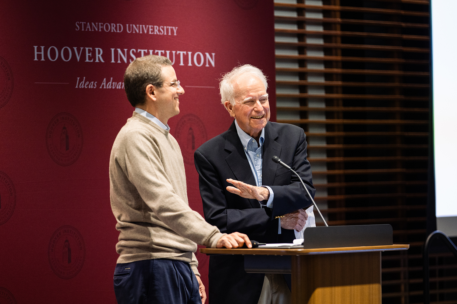 Robert Hall speaking at a podium, with a smiling man standing beside him in front of a Hoover Institution banner.