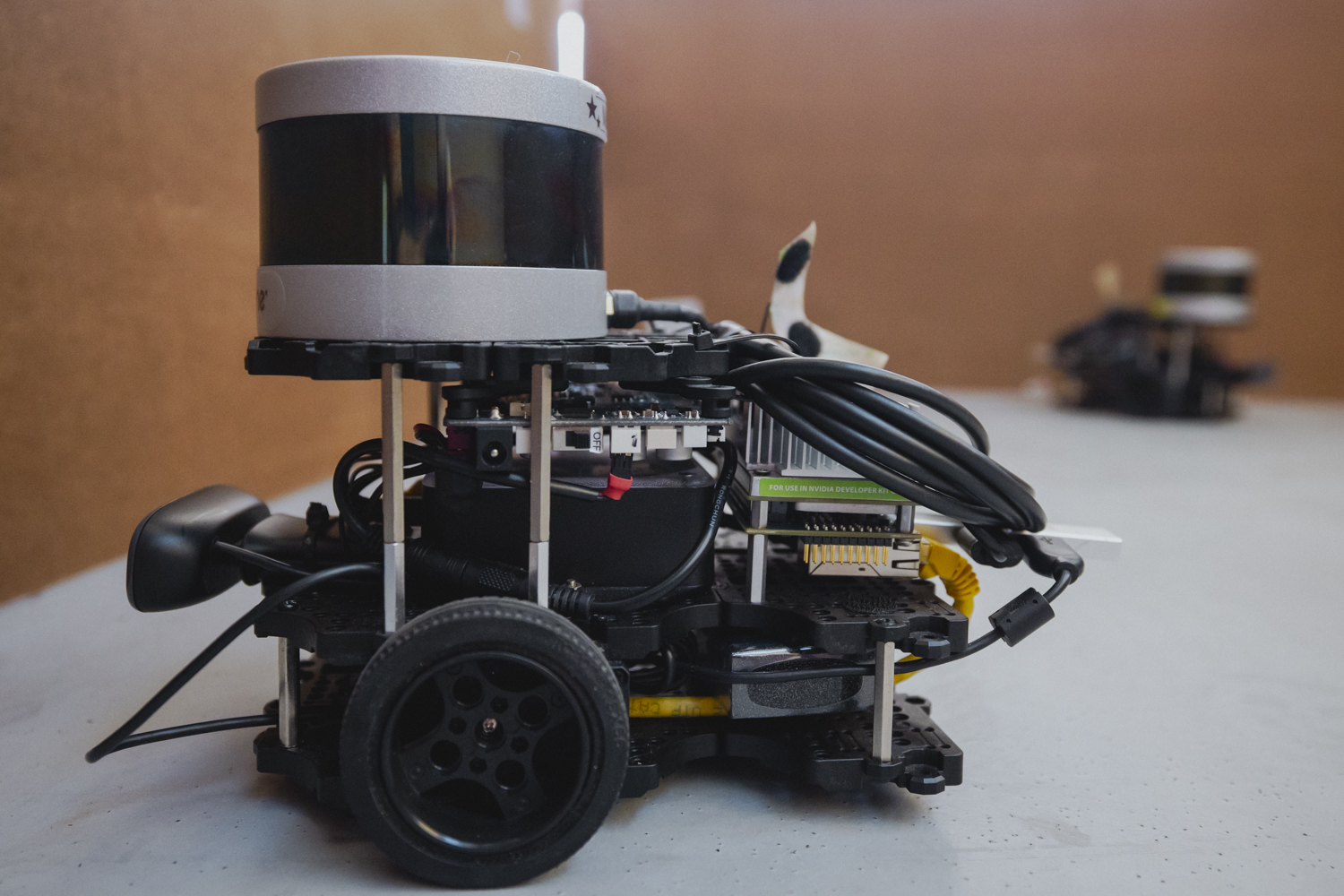 Small wheeled robot with a cylindrical lidar sensor, circuit boards and cables on a table, another robot blurred behind.