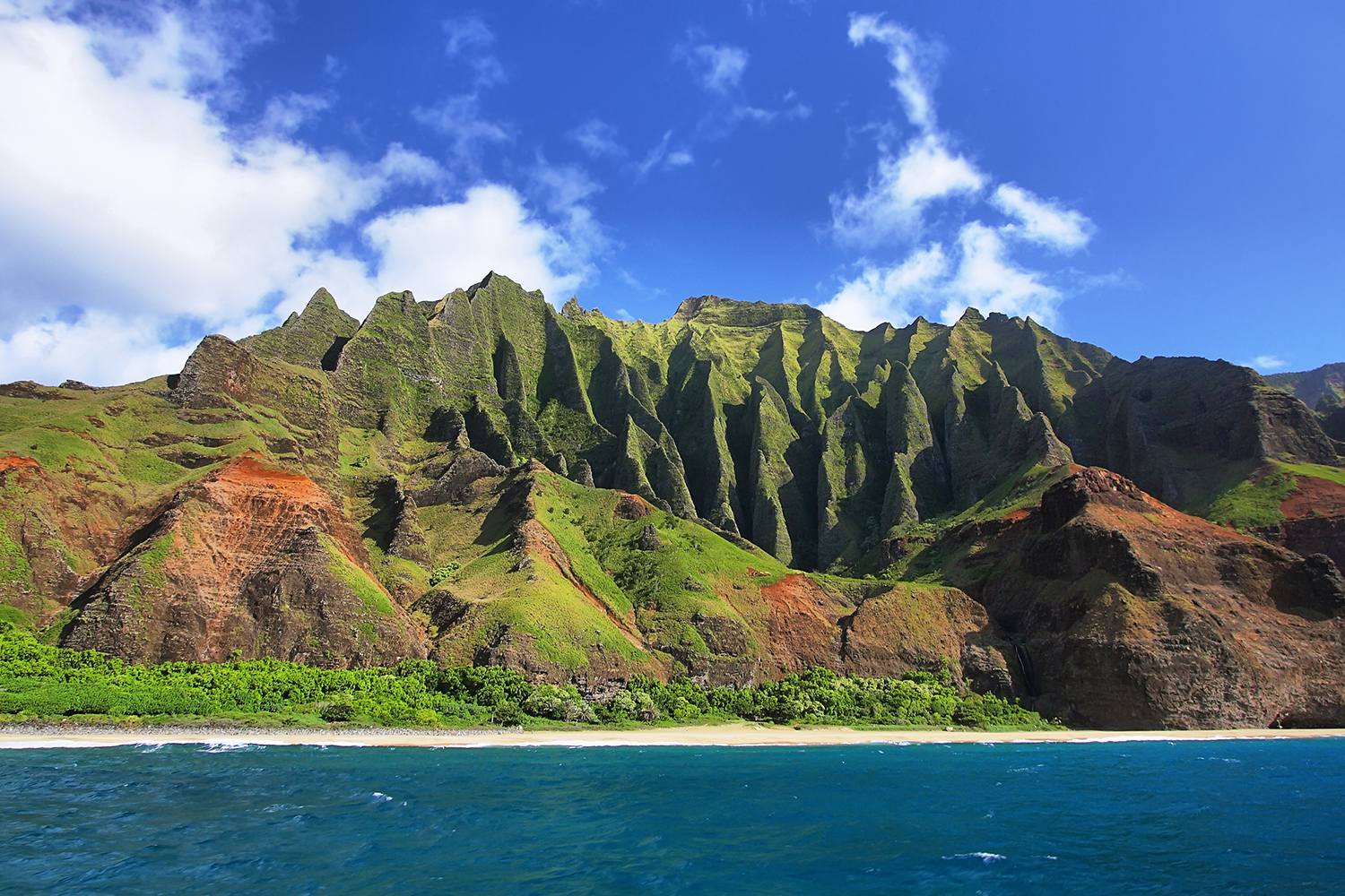 Hawaii's Na Pali coast with steep green volcanic ridges and red cliffs rising above a sandy beach and deep blue ocean under a partly cloudy sky.