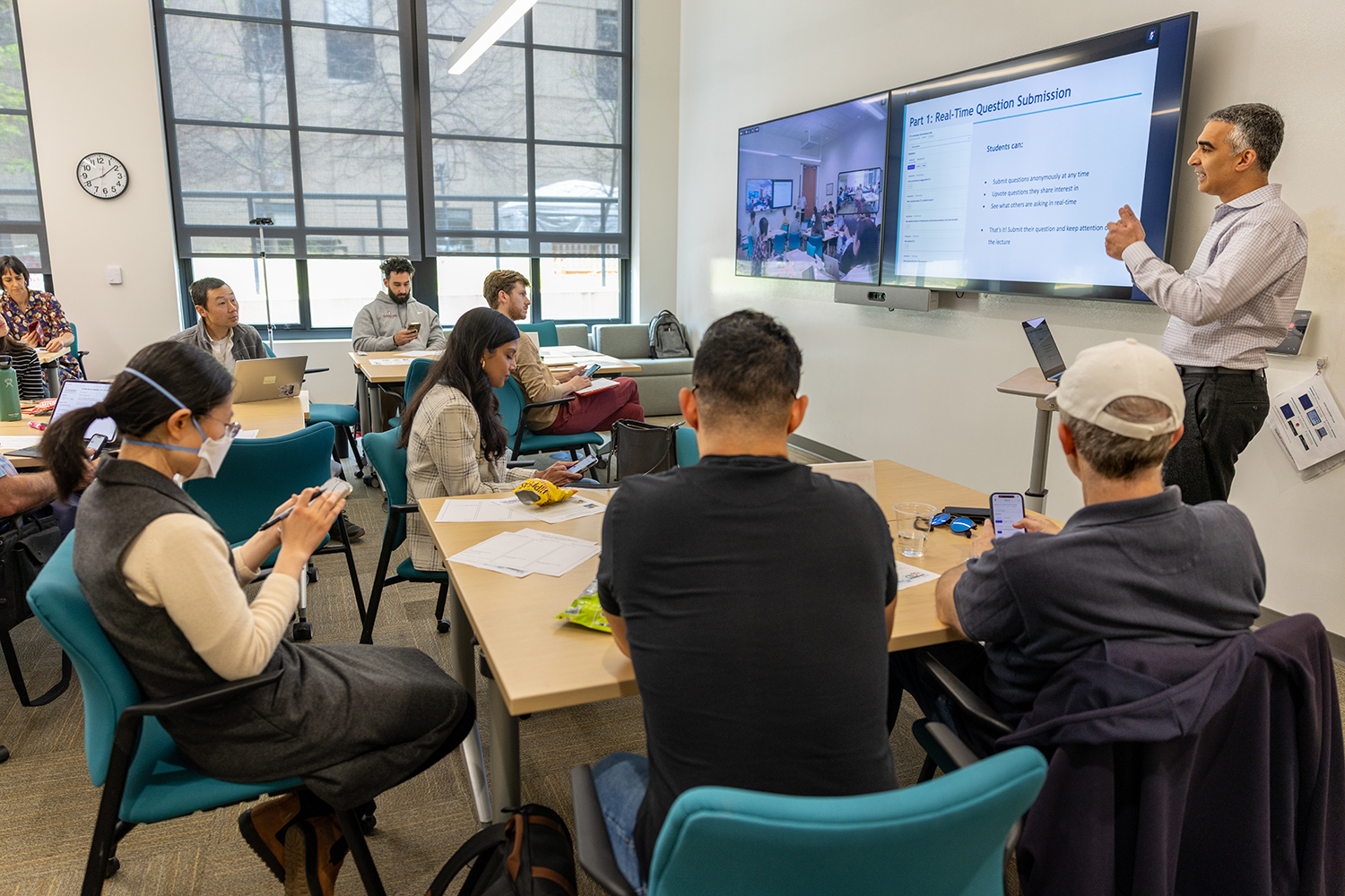 Amit Kaushal gestures at a screen showing “Real-Time Question Submission” while workshop participants use their phones at tables.