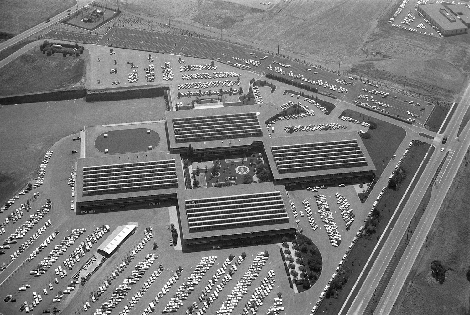 Black-and-white aerial view of the research park complex in the 1960s with four long wings around a courtyard, surrounded by parking.