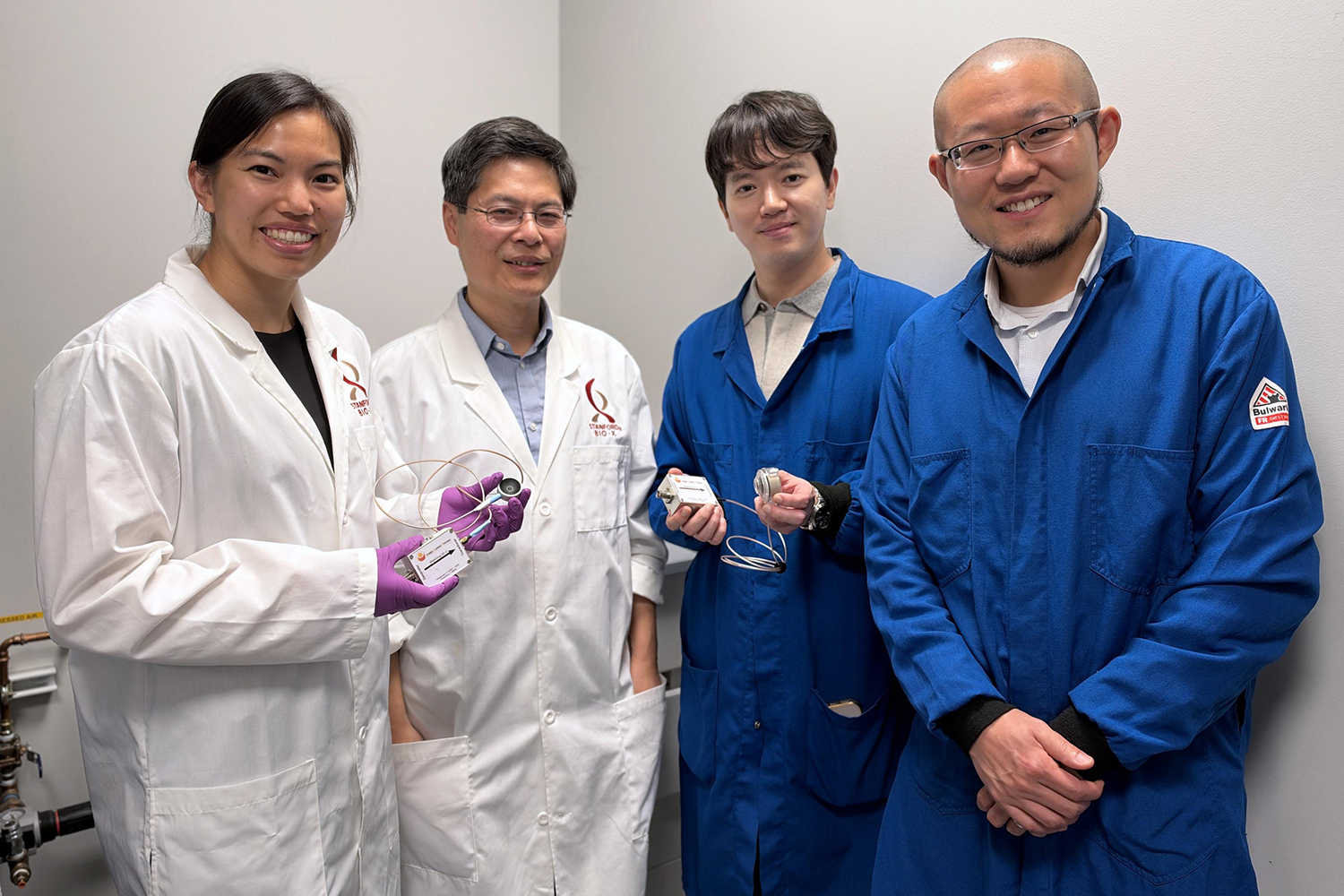 Four researchers stand together, two holding small wired sensor devices, showcasing a lab technology project while wearing white and blue lab coats.