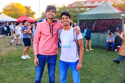 Two friends smiling in the foreground, festival booths in the background.