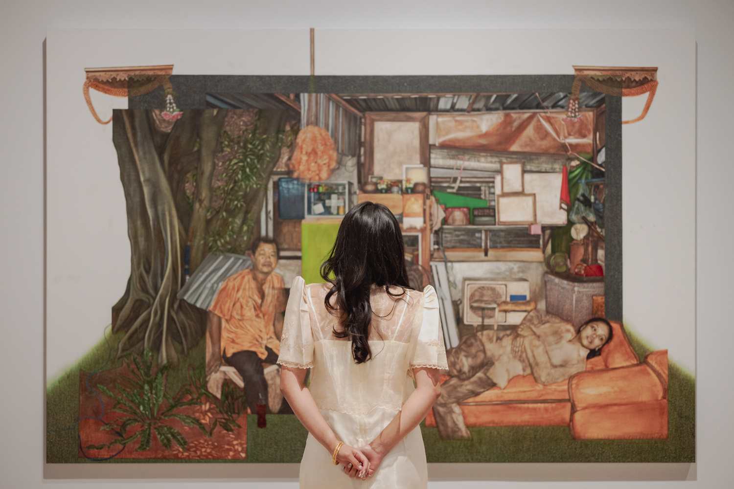 A person considers a large painting. The painting depicts a small, rectangular house, hemmed in on one side by tropical trees, and its two inhabitants. The house is full of colorful items and appears to be spilling out onto the grass. The scene is presented almost as a diorama or a shrine.