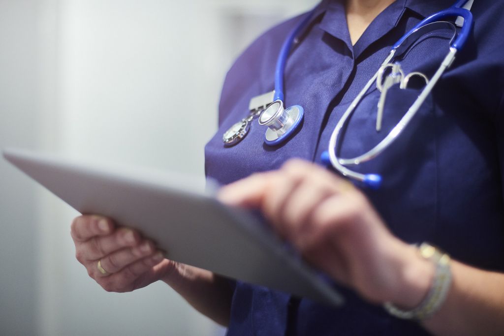 Nurse wearing a dark blue top and stethoscope around her neck is looking at patient's record on her digital tablet.