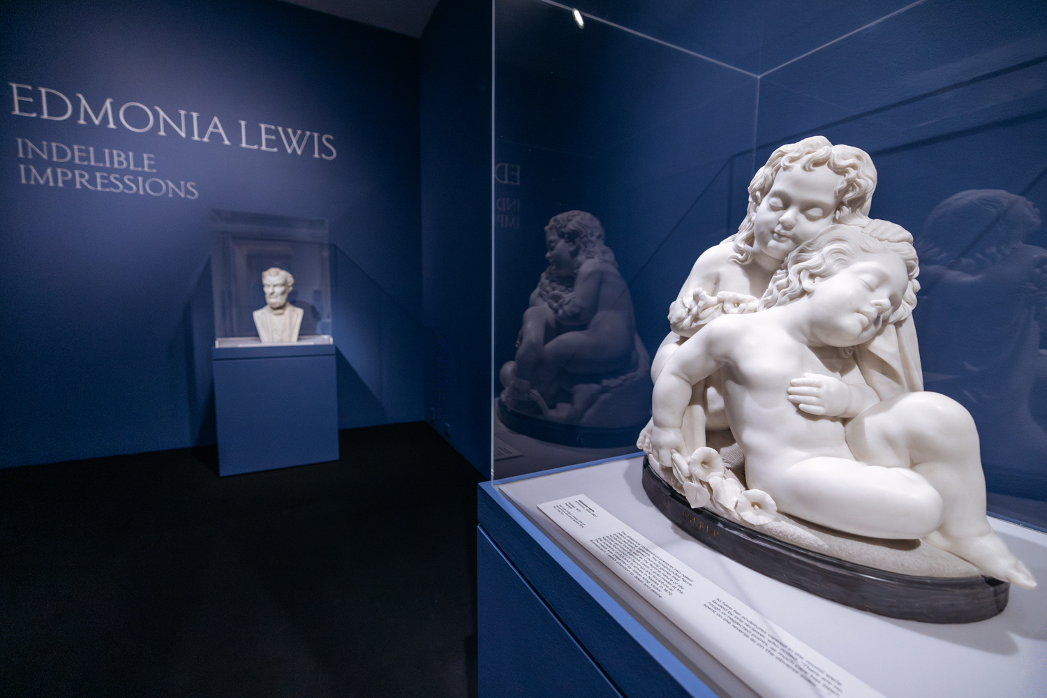A marble sculpture depicting two seated people is displayed in a blue-walled gallery labeled 'Edmonia Lewis: Indelible Impressions.' A third sculpture, a bust, is visible in the background.