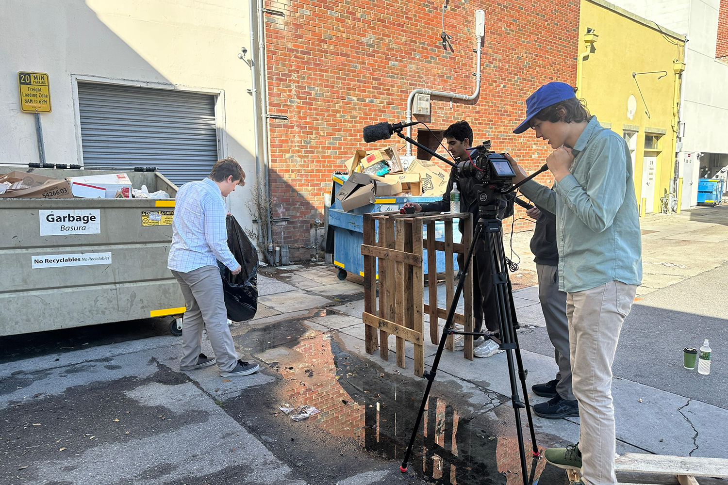 A filming crew captures footage in an alley, featuring a dumpster and a person holding a garbage bag.