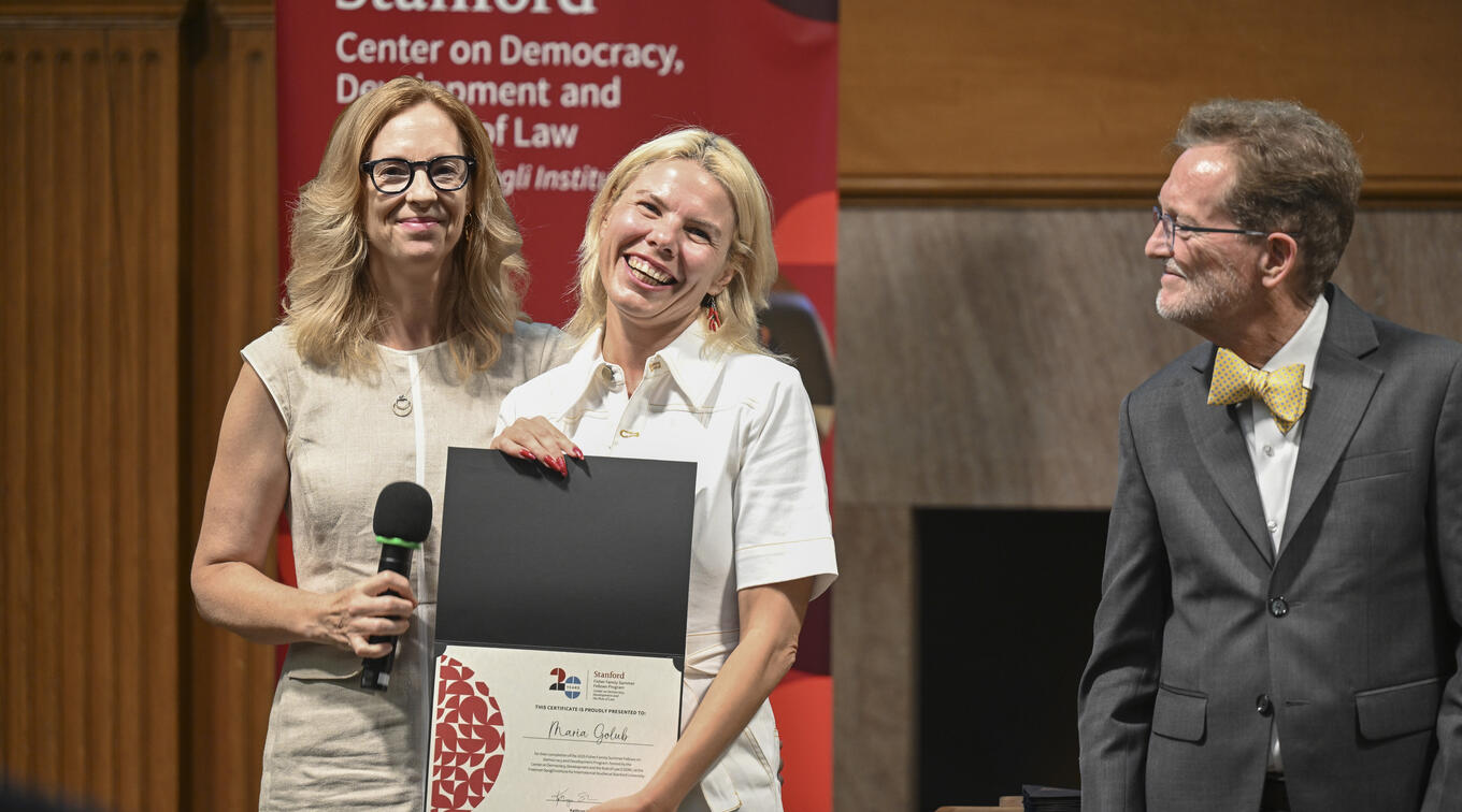 Maria Golub and another woman stand together on stage, Golub holding a certificate, while an older man in a suit looks on during an awards ceremony.