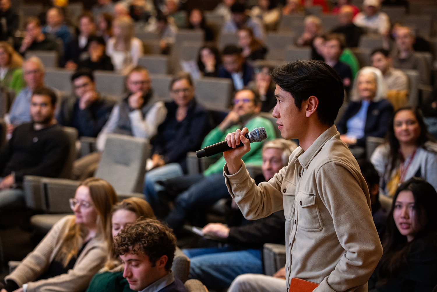A participant asking a question into a microphone during a crowded event, while seated attendees listen attentively.