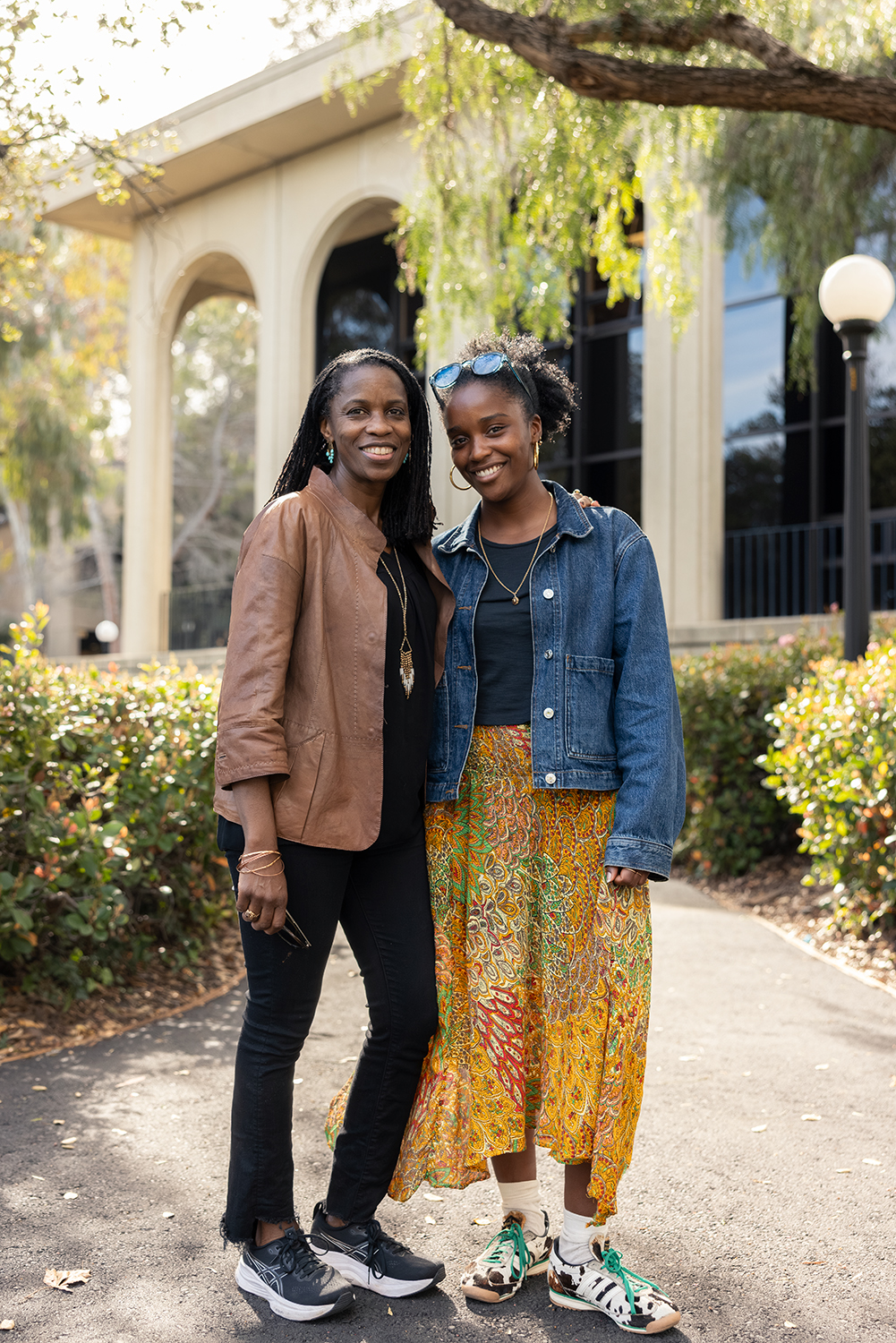 Two women pose together outdoors, one in a brown jacket and the other in a denim jacket and colorful skirt, against a leafy backdrop.